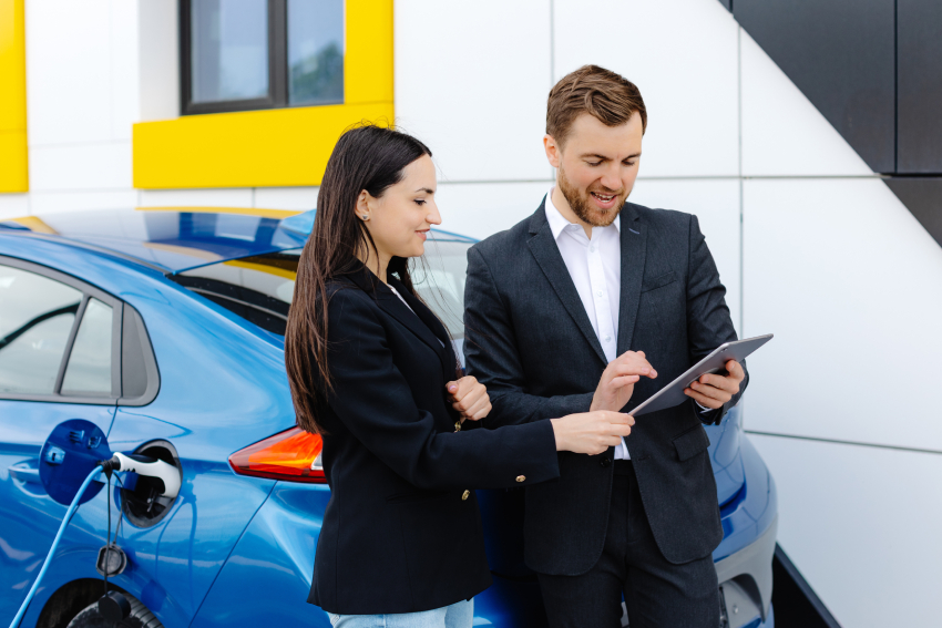 Smiling woman standing at the car and pointing at car seller tablet. She is picked right car for her she saw online. A woman concludes an agreement to buy a car and shakes the manager's hand